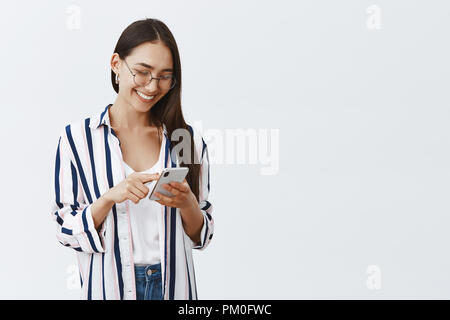 Choosing new wallpaper on smartphone. Portrait of happy attractive and stylish woman in glasses and striped blouse scrolling feed while using smartphone, reading amusing article in internet Stock Photo