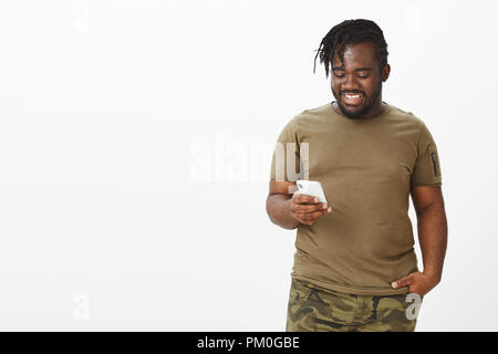 Indoor shot of good-looking happy dark-skinned guy in military outfit, holding smartphone and looking at screen with satisfied expression, being joyful to receive good news via internet Stock Photo