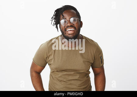 Smile keeps sadness away. Portrait of bright good-looking african american male model in glasses and military t-shirt, laughing out loud and expressing joyful happy attitude over grey wall Stock Photo