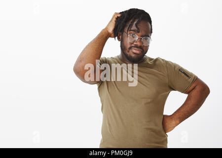 Indoor shot of unsure handsome african american in glasses, holding hand on hip and scratching head, being doubtful while thinking, making choice with confused expression, standing in hesitations Stock Photo