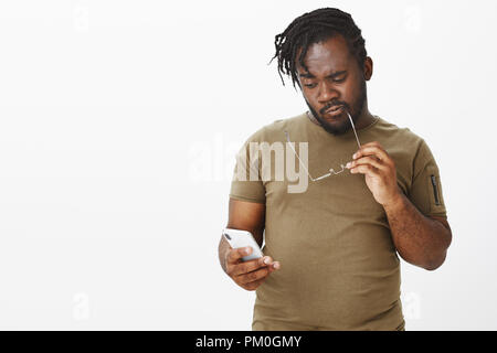 Studio shot of thoughtful smart african man in casual outfit, biting rim of glasses while holding smartphone and gazing at screen, thinking, making decision or choice while standing over grey wall Stock Photo