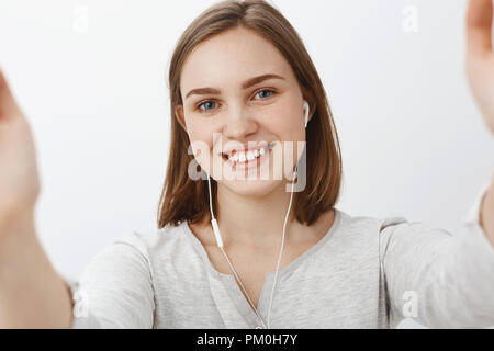 Friendly looking cute and happy young woman with short brown haircut smiling joyfully wearing earphones and pulling hands towards camera as if taking selfie or talking via video message in smartphone Stock Photo