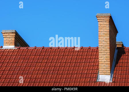 Fragment of the roof of the red metal tiles with chimneys of beige brick, against the bright blue sky. Stock Photo