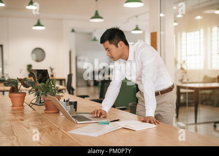 Focused young Asian businessman leaning on his desk in a modern office working online with a laptop and going over paperwork Stock Photo