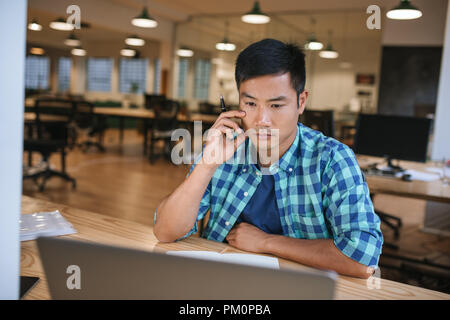 Focused young Asian designer sitting at his workstation using a laptop while working late in a modern office Stock Photo
