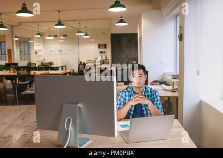 Portrait of a focused young Asian designer working with a laptop and monitor while sitting alone at his desk in a modern office Stock Photo