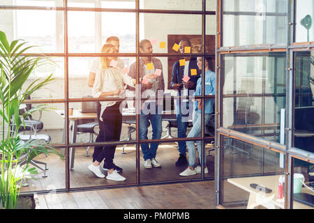 Sharing business ideas. Full length of young modern people in smart casual wear using adhesive notes while standing behind the glass wall in the board room. Stock Photo