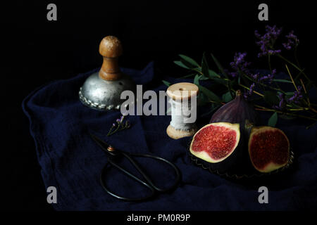 Dark still life with ripe fresh purple figs on linen tablecloth. Vintage moody composition with fruit, old scissors, silk ribbon, eucalyptus branches and flowers. Selective focus. Stock Photo