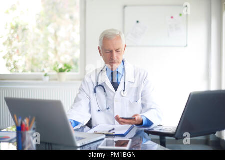 Elderly male doctor using his mobile phone and working on laptop while sitting at private clinic. Stock Photo