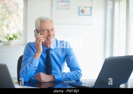 Portrait of senior investment advisor businessman wearing shirt and tie and talking with somebody on his mobile phone while working on laptops in the  Stock Photo