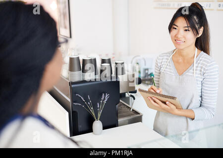 Young female in workwear talking to client and writing down order in notepad during work Stock Photo