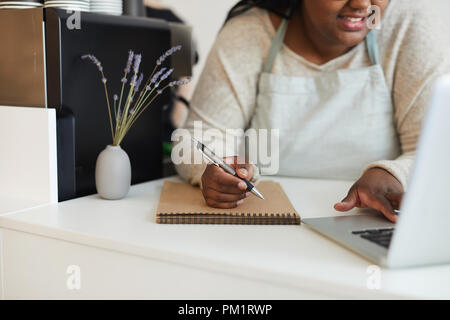 Modern waitress in apron leaning over counter while browsing in the net and making notes Stock Photo