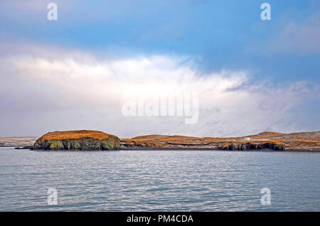 View across Reykjavik bay to the grassy hills and rocky cliffs of Videy island and the mountains beyond under a sky with low hanging clouds Stock Photo