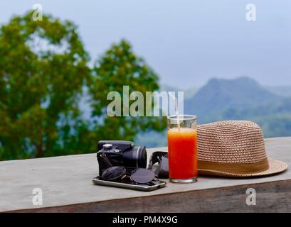 Close up of travel essentials, sunhat, camera, mobile and sunglasses against background of beautiful mountainous scenery with ample editorial space Stock Photo