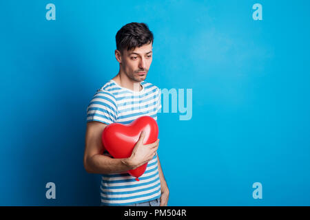 Portrait of a sad young man in a studio on a blue background, holding red heart. Stock Photo