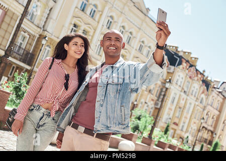 Romantic Relationship. Young diverse couple walking on the city street holding hands taking selfie photo on smartphone smiling cheerful Stock Photo