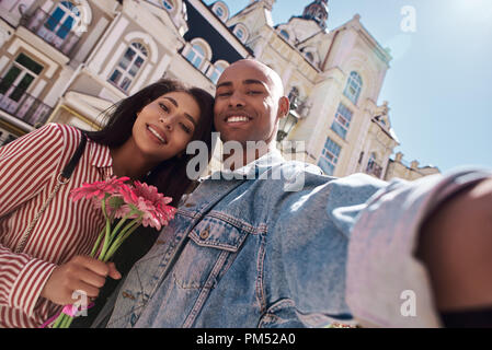 Romantic Relationship. Young diverse couple walking on the city street girl holding flowers while guy taking  selfie on smartphone smiling happy close-up Stock Photo