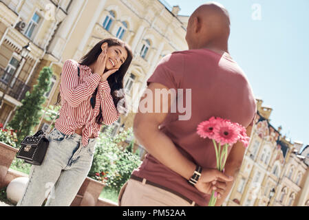 Romantic Surprise. Young diverse couple standing on the city street boyfriend holding bouquet of flowers behind back while girlfriend looking at him smiling cheerful Stock Photo