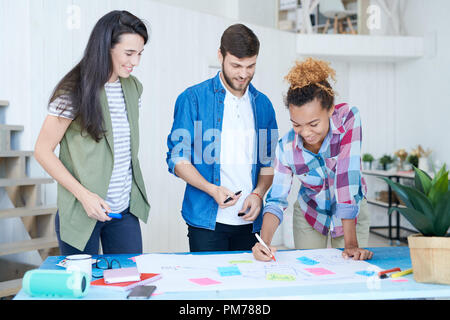 Multi-ethnic group of contemporary young people drawing charts and graphs while planning startup project standing at desk and smiling happily Stock Photo