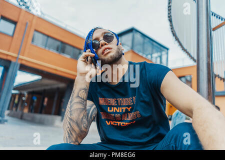 Young attractive man with blue dreadlocks talking by mobile phone and touching his hair. Man is on foreground and focus, red bricked building and art  Stock Photo