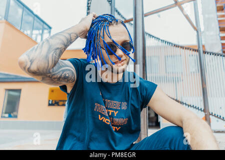 Young attractive man with blue dreadlocks touching his hair, and sitting next to art object. Stock Photo