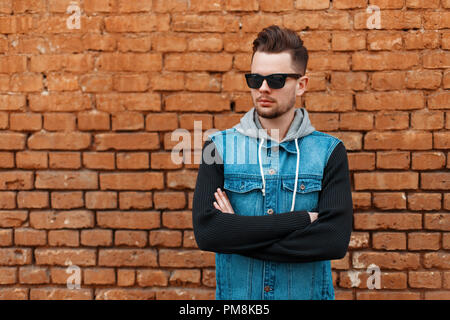 Handsome man in sunglasses and a denim waistcoat near a brick wall Stock Photo