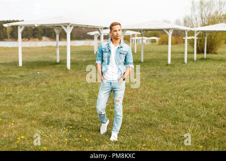 Handsome man in stylish denim clothing and a white T-shirt, sneakers walking on the beach with white canopies Stock Photo