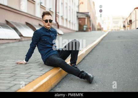Handsome young man in a blue shirt, black jeans and sneakers sits on the sidewalk Stock Photo