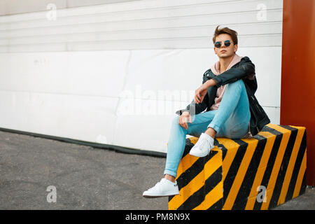 Stylish handsome young man in a black leather jacket, a pink sweatshirt and white sneakers sits on a black-and-yellow concrete Stock Photo