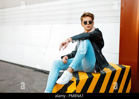 Fashionable handsome young man in branded stylish clothes sits on black-and-yellow concrete Stock Photo
