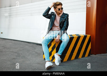 Handsome young model of a man wearing sunglasses in a black leather jacket, a pink sweatshirt, blue jeans and white shoes sits on a black-and-yellow s Stock Photo