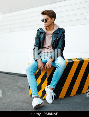 Young handsome man with sunglasses in a stylish black leather jacket, a pink sweatshirt, blue jeans and white shoes sits on a black-and-yellow slab Stock Photo