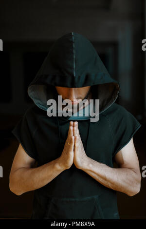 Sporty yoga man in a hood holds his hands on a dark background Stock Photo