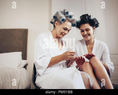 Two women in bathrobe with curly rollers pinned on hair sitting together at home. Mother applying nail polish to her daughter. Stock Photo