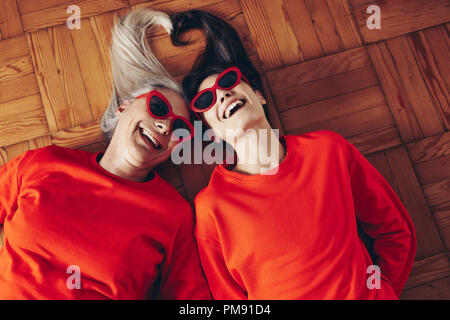 Two happy women in fancy sunglasses and orange t shirts lying on floor. Women lying on floor together making a heart shape with their hair. Stock Photo