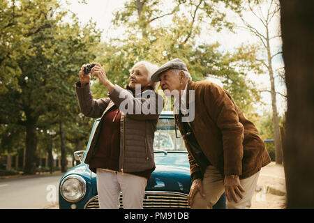 Senior couple on road trip taking pictures of nature landscape with digital camera. Old woman taking photographs with her camera with man standing by Stock Photo