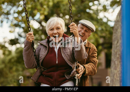 Senior man pushing his female partner on swing in park and having fun together. Playful and happy senior couple enjoying at swing in park . Stock Photo