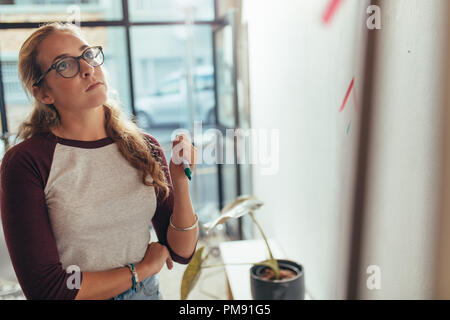 Young woman standing in office and looking at the presentation board thinking. Female computer programmer thinking over new project plan at tech start Stock Photo