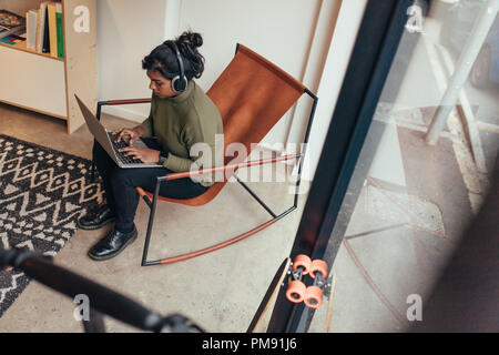 Young woman wearing headphones sitting on chair and working on laptop computer. Female computer programmer working at her startup office. Stock Photo