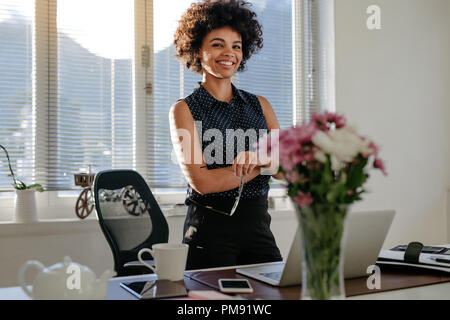 Business woman standing next to her desk in office. Confident african woman with her arms crossed wearing casual attire at her workplace.