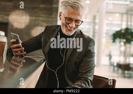 Smiling mature male in business suit sitting at cafe in earphones and holding a mobile phone. Business man making phone call using earphones in coffee Stock Photo