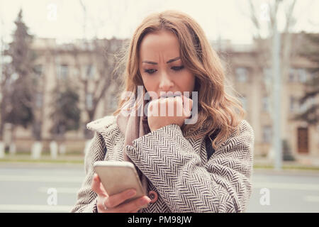 Young nervous woman watching smartphone and biting fist while standing on street Stock Photo