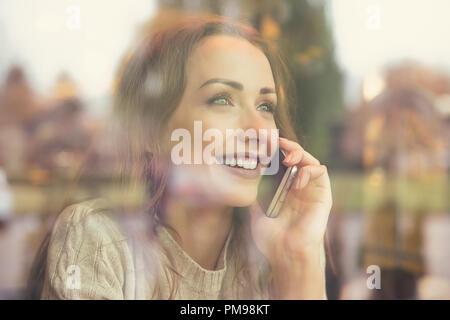 View through window of coffee shop of young happy woman having phone call and smiling away Stock Photo