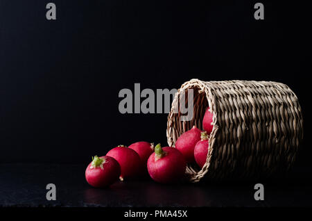 Basket filled with radishes on its side and spilling out on to slate surface with black background. Stock Photo