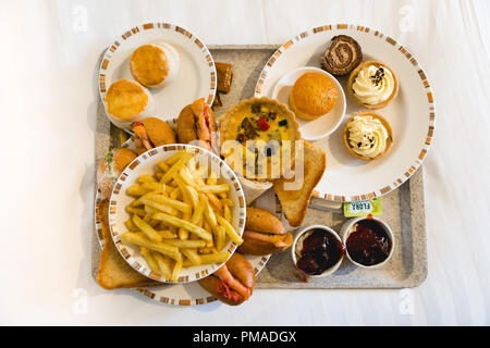 Tray full of sweet and savoury food including plates of sandwiches, rolls, cakes, pastries, scones & French fries (chips) for afternoon tea, on a bed