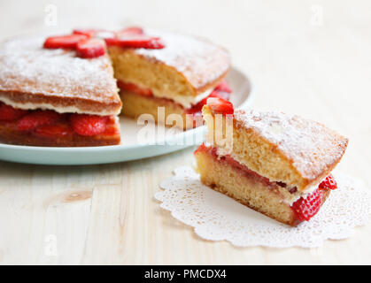Victoria sponge cake with strawberries, jam and whipped cream with a cut out piece on a wooden table Stock Photo