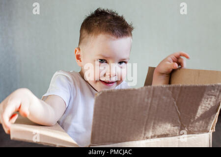 a 4 year old blond boy sits in a cardboard box at home and plays hide and seek, pick a boo Stock Photo