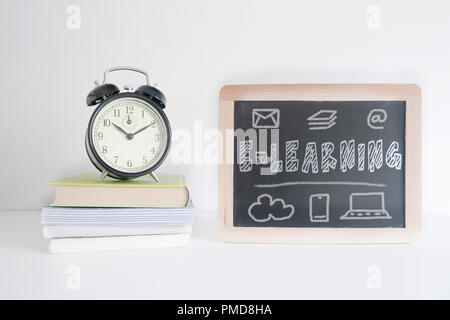Alarm clock on a pile of books next to a blackboard with the text E-LEARNING written on it. Empty copy space for Editor's text. Stock Photo