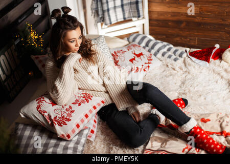 Beautiful fashionable young girl with a haircut in a knitted vintage sweater with red socks on the bed with Christmas decorations Stock Photo