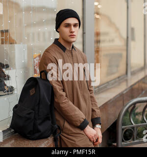Fashionable young man in a stylish vintage coat with a hat and a black backpack sitting near the shop window on the street Stock Photo